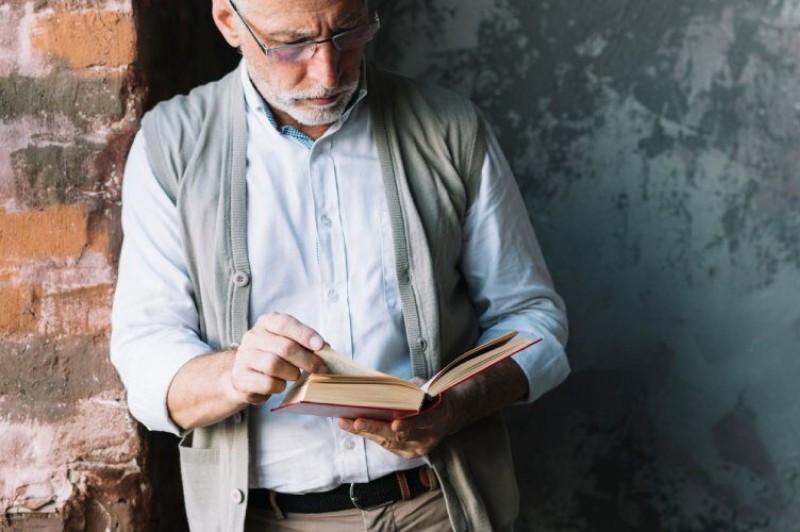 hombre adulto leyedo un libro de crecimiento personal, con pelo y barba blanca, vestimenta informal, apoyado sobre una columna
