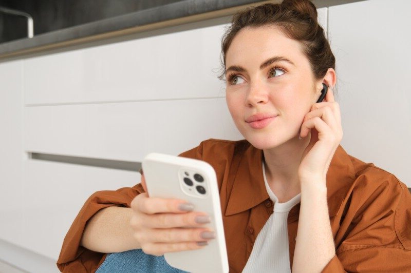 Chica joven, morena, con camisa marrón, escuchando podcasts de crecimeinto personal con los auriculares en el teléfono móvil